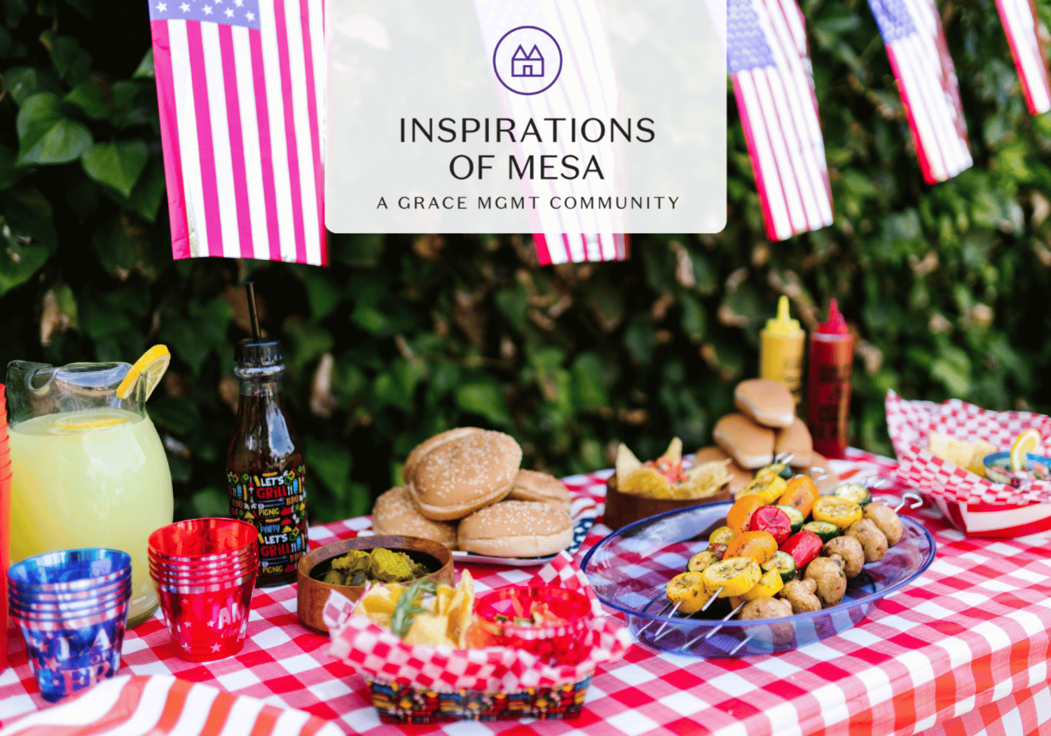 Patriotic outdoor picnic table with burgers, skewers, lemonade, and sauces on a red-and-white checkered cloth under American flags and a sign reading 'Inspirations of Mesa'.